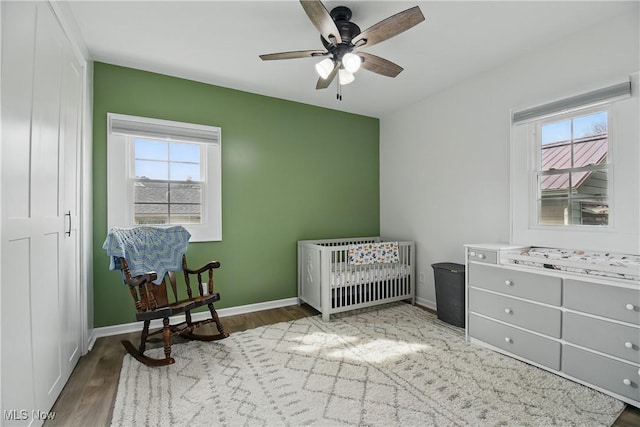 bedroom featuring a crib, wood finished floors, baseboards, and ceiling fan