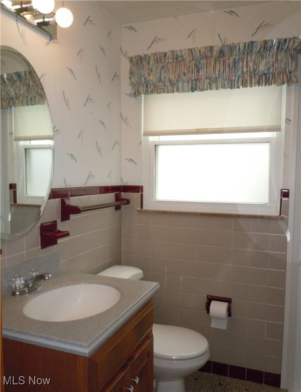 half bath featuring toilet, tile walls, wainscoting, wallpapered walls, and vanity