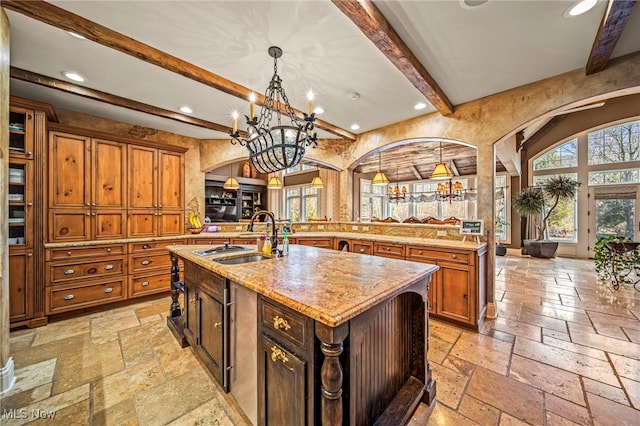 kitchen featuring brown cabinets, an island with sink, a sink, stone tile floors, and arched walkways