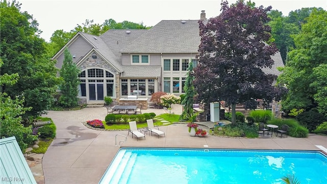 back of property featuring stone siding, a patio, roof with shingles, an outdoor pool, and a chimney