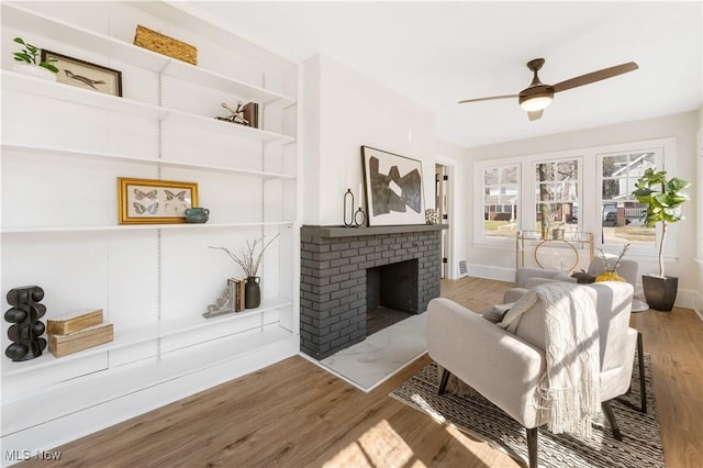 living area featuring a ceiling fan, built in features, wood finished floors, baseboards, and a brick fireplace