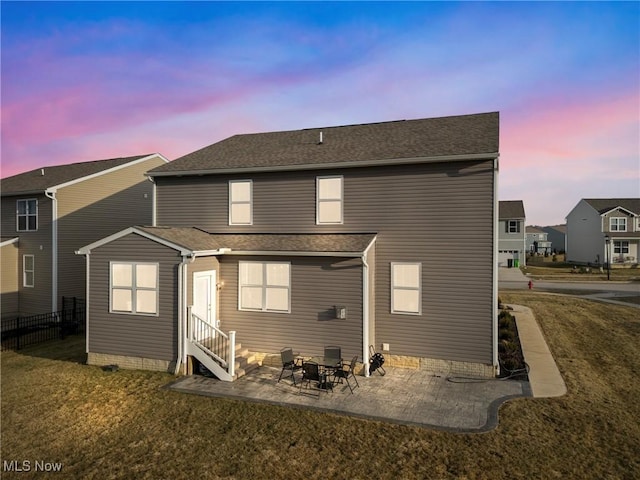 back of property at dusk featuring a shingled roof, fence, entry steps, a lawn, and a patio area