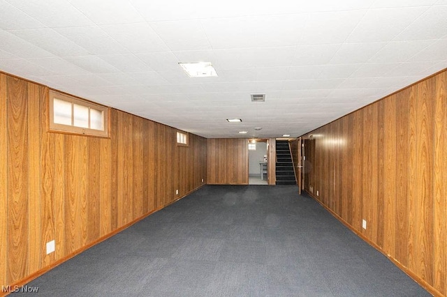 basement with wooden walls, dark carpet, stairway, and visible vents