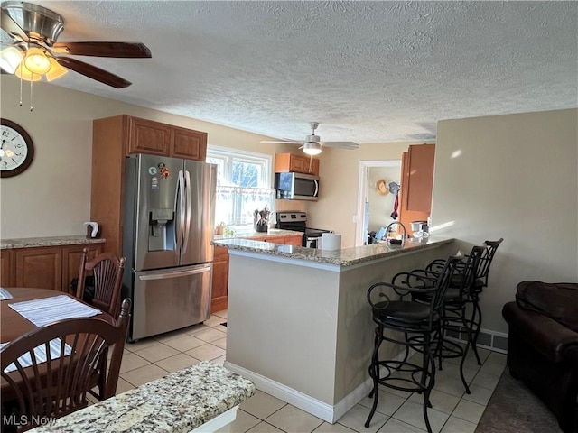 kitchen featuring light tile patterned floors, a ceiling fan, a peninsula, stainless steel appliances, and brown cabinets