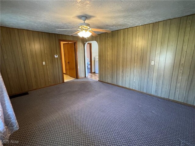 carpeted empty room featuring wooden walls, a ceiling fan, arched walkways, and a textured ceiling