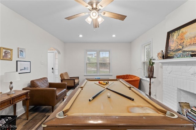 recreation room with a ceiling fan, dark wood-type flooring, a brick fireplace, arched walkways, and recessed lighting