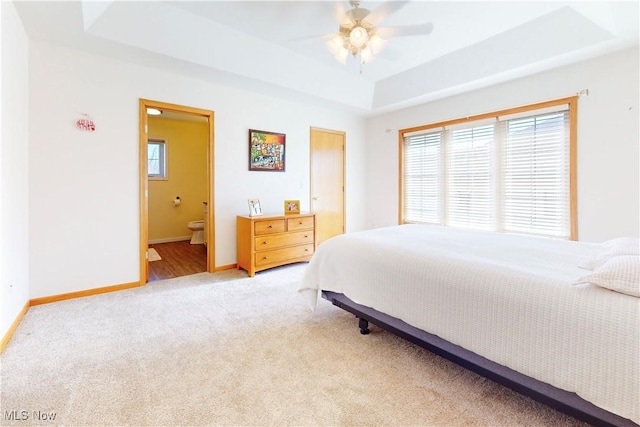 bedroom with baseboards, light colored carpet, a tray ceiling, ensuite bathroom, and a ceiling fan