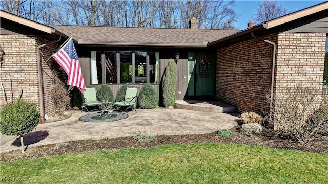 doorway to property featuring a patio, brick siding, a chimney, and roof with shingles