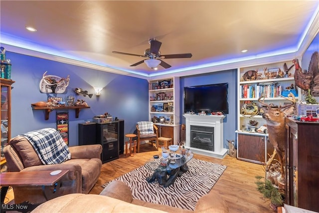 living area featuring a tray ceiling, built in shelves, wood finished floors, a glass covered fireplace, and ceiling fan