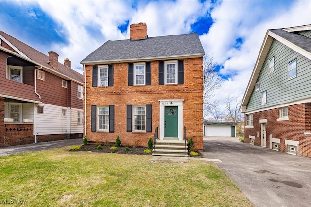 colonial house with a front yard, roof with shingles, a chimney, an outbuilding, and brick siding