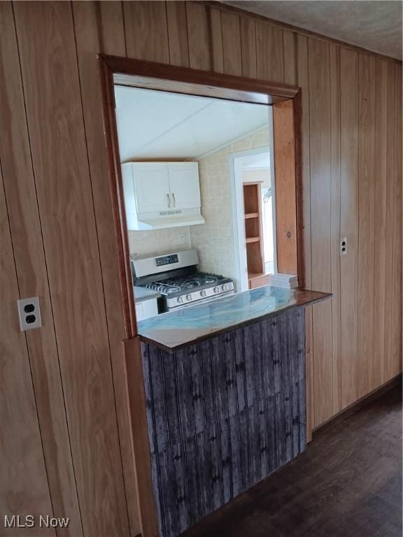 kitchen with wooden walls, stainless steel range with gas stovetop, under cabinet range hood, white cabinetry, and dark wood-style floors