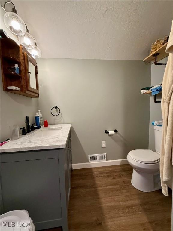 half bath with vanity, toilet, a textured ceiling, visible vents, and wood finished floors