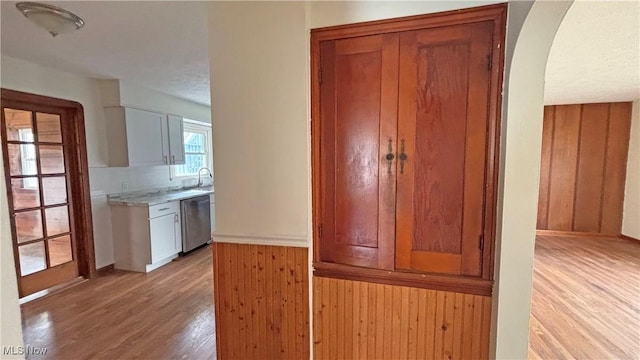 kitchen with light countertops, stainless steel dishwasher, arched walkways, wooden walls, and light wood finished floors