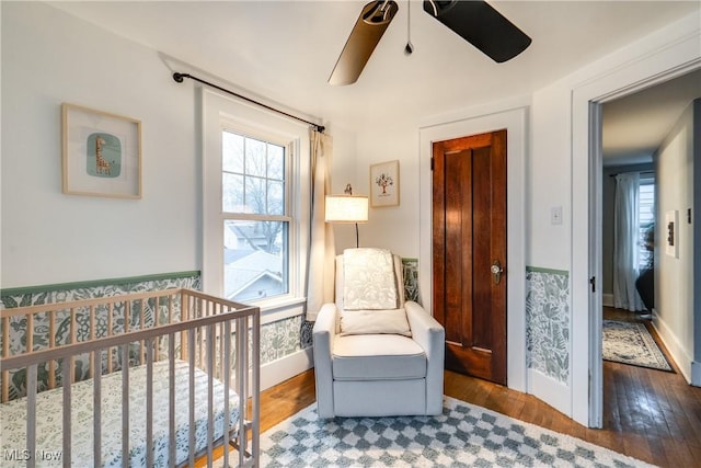 bedroom with wainscoting, a crib, a ceiling fan, and wood finished floors