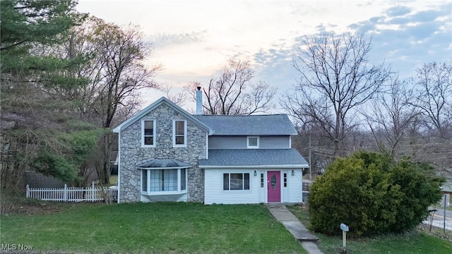 traditional home featuring a front yard, stone siding, a chimney, fence, and roof with shingles