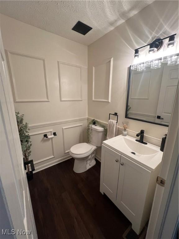 half bathroom featuring a decorative wall, a textured ceiling, vanity, toilet, and wood finished floors