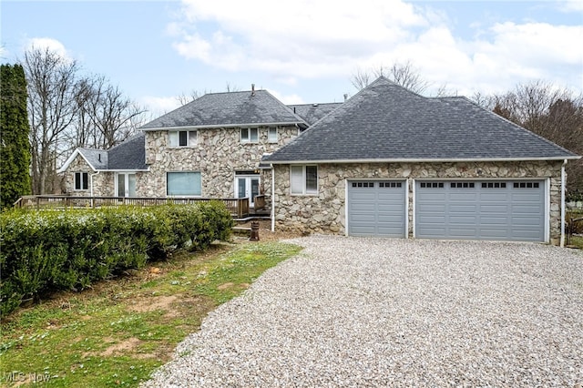 view of front facade with gravel driveway, stone siding, a garage, a wooden deck, and roof with shingles