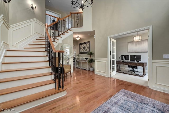 stairway with a high ceiling, a decorative wall, a chandelier, wood finished floors, and a wainscoted wall