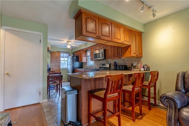 kitchen with a peninsula, a ceiling fan, a breakfast bar, appliances with stainless steel finishes, and brown cabinets