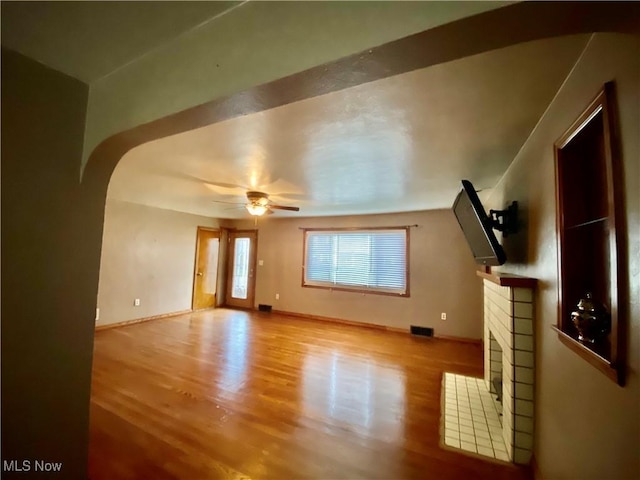 unfurnished living room featuring baseboards, a fireplace, ceiling fan, arched walkways, and wood finished floors
