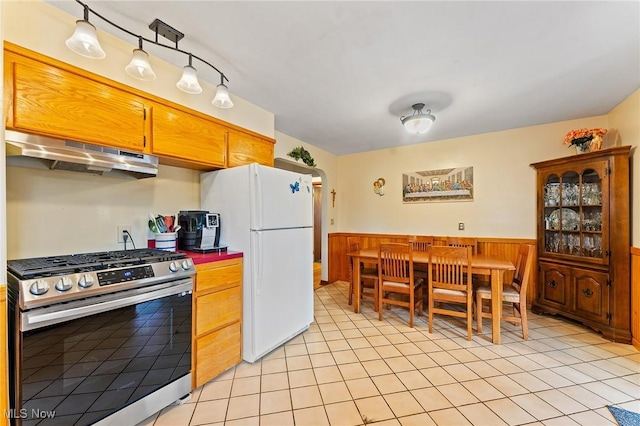 kitchen featuring a wainscoted wall, stainless steel gas stove, under cabinet range hood, light tile patterned floors, and freestanding refrigerator