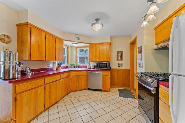 kitchen with under cabinet range hood, wooden walls, a sink, wainscoting, and appliances with stainless steel finishes