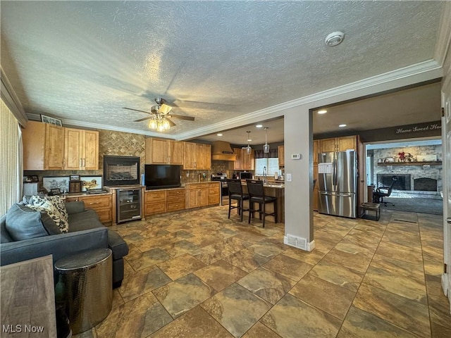 living area featuring wine cooler, a ceiling fan, crown molding, a fireplace, and a textured ceiling