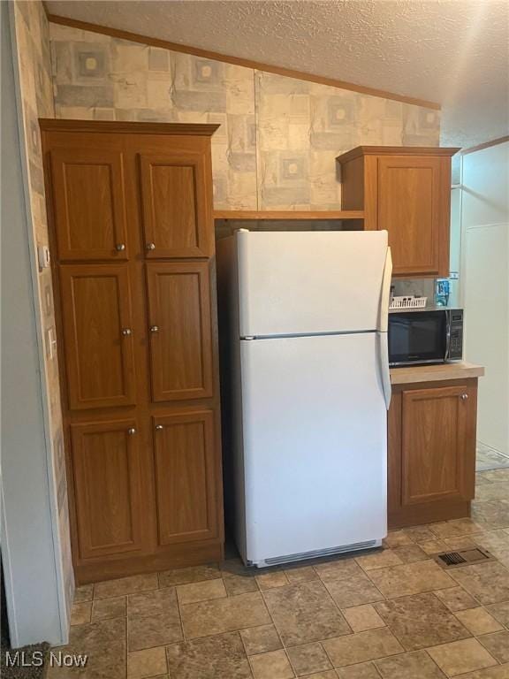 kitchen featuring a textured ceiling, wallpapered walls, black microwave, stone finish floor, and freestanding refrigerator