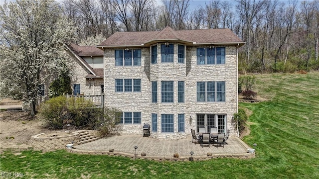 back of house featuring a lawn, stone siding, a patio, and a shingled roof