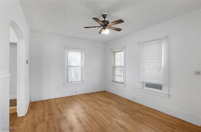 spare room featuring a ceiling fan, arched walkways, baseboards, light wood-style flooring, and a textured ceiling
