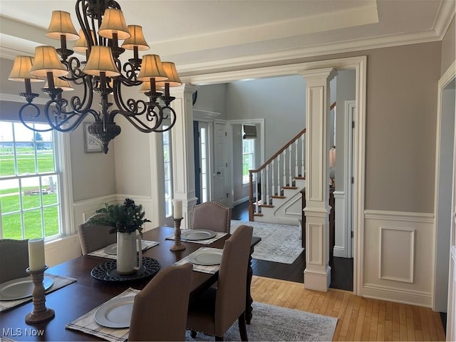 dining space featuring a wainscoted wall, decorative columns, light wood-style floors, a chandelier, and stairs
