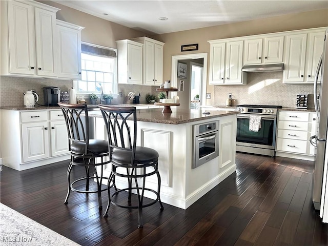 kitchen with under cabinet range hood, stainless steel appliances, backsplash, a kitchen island, and white cabinetry