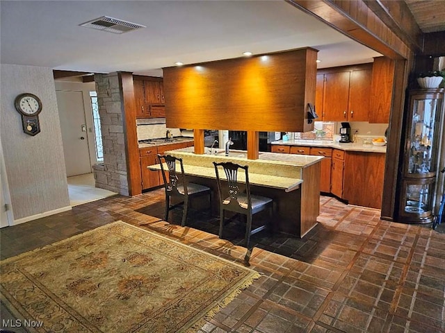 kitchen with visible vents, light countertops, baseboards, an island with sink, and brown cabinetry