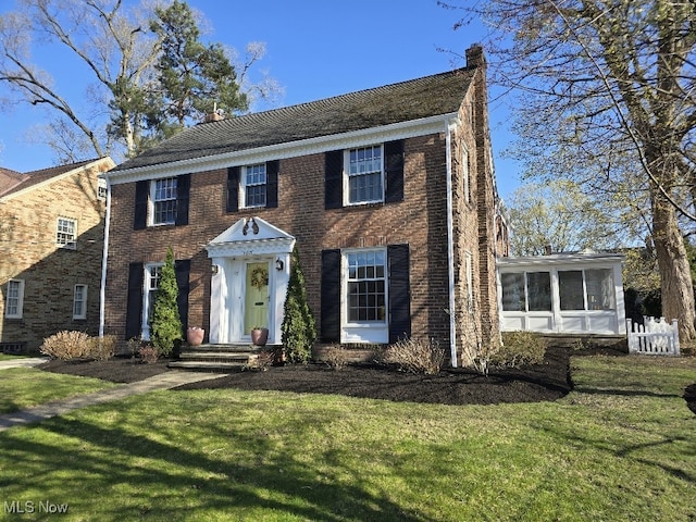 colonial inspired home featuring brick siding, a front yard, a sunroom, and a chimney