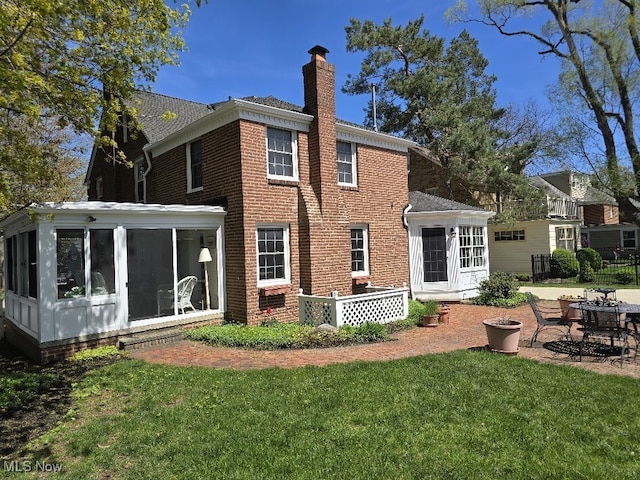 back of house featuring a chimney, brick siding, a patio area, a lawn, and a sunroom