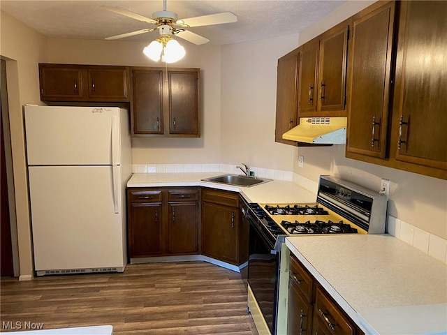 kitchen with a sink, freestanding refrigerator, gas range oven, under cabinet range hood, and dark wood-type flooring