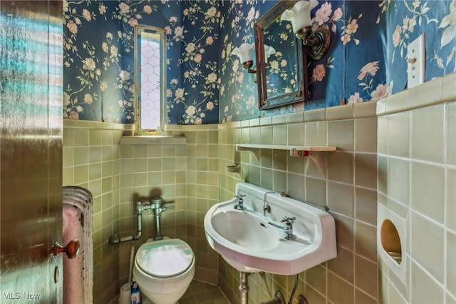 half bathroom featuring a sink, wallpapered walls, wainscoting, tile walls, and toilet
