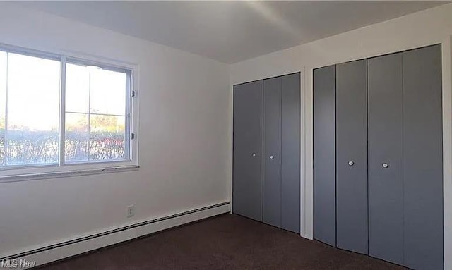 unfurnished bedroom featuring a baseboard heating unit, two closets, and dark carpet