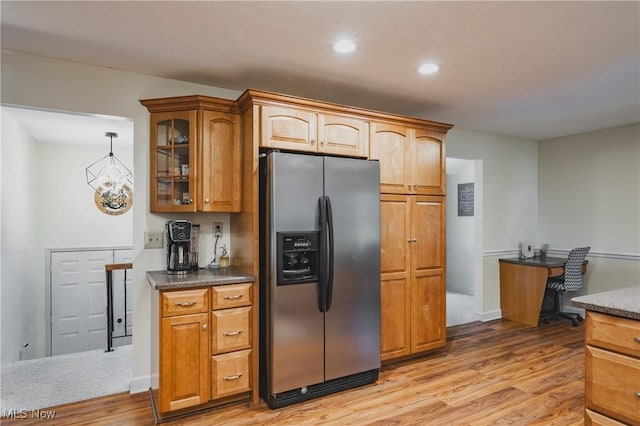 kitchen with glass insert cabinets, stainless steel fridge, brown cabinetry, light wood-style flooring, and dark countertops
