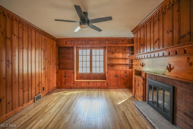 unfurnished living room with wood walls, visible vents, light wood finished floors, a brick fireplace, and a ceiling fan