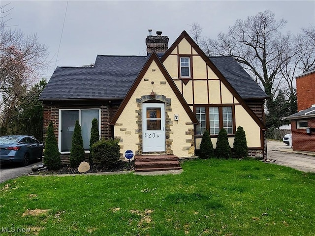 tudor house with a front lawn, a shingled roof, stucco siding, and a chimney