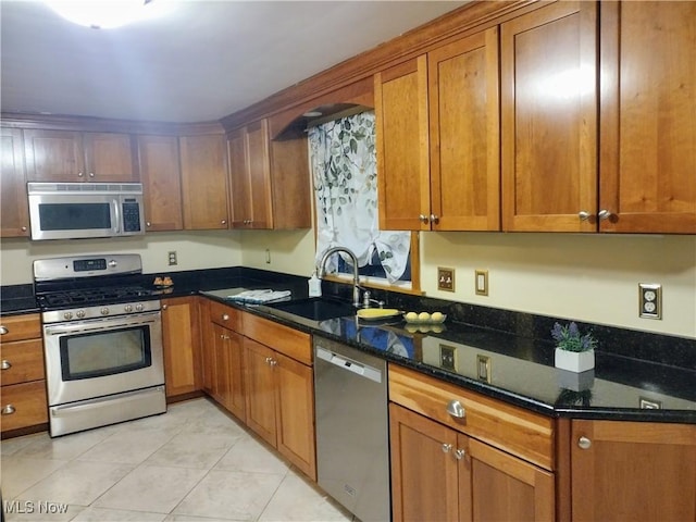 kitchen featuring dark stone countertops, a sink, appliances with stainless steel finishes, light tile patterned flooring, and brown cabinetry