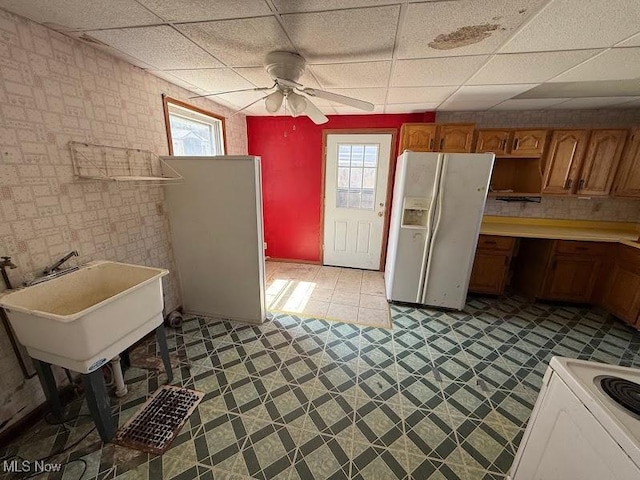 kitchen with white refrigerator with ice dispenser, ceiling fan, electric range oven, a paneled ceiling, and brown cabinetry