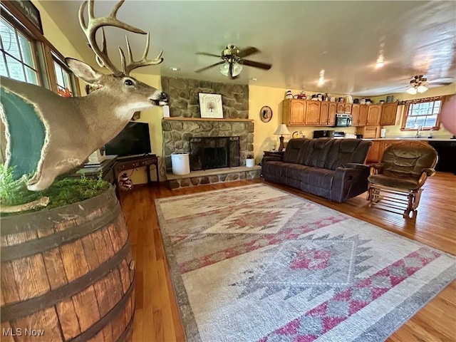 living room featuring a ceiling fan, a stone fireplace, and wood finished floors