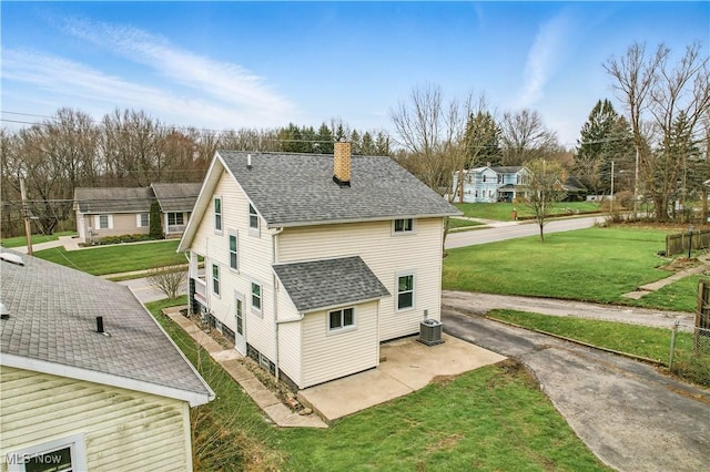 view of side of property with a patio area, a chimney, cooling unit, a lawn, and roof with shingles