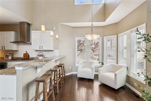 kitchen featuring wall chimney exhaust hood, light stone counters, a towering ceiling, and white cabinetry