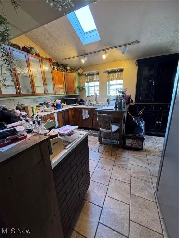 kitchen with a textured ceiling, stainless steel microwave, brown cabinets, glass insert cabinets, and rail lighting