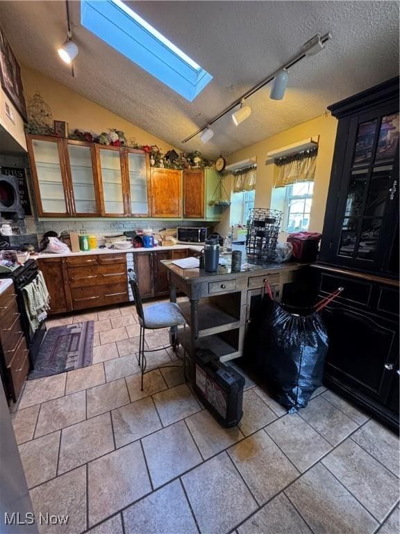 kitchen featuring stainless steel microwave, brown cabinets, light countertops, rail lighting, and lofted ceiling with skylight