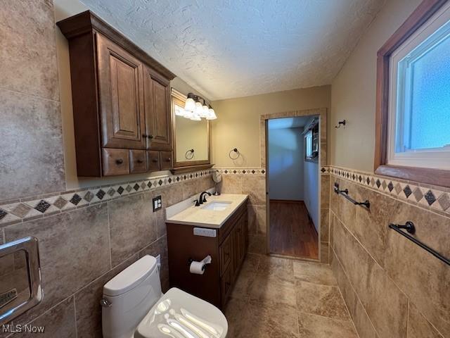 bathroom with vanity, toilet, a textured ceiling, a wainscoted wall, and tile walls