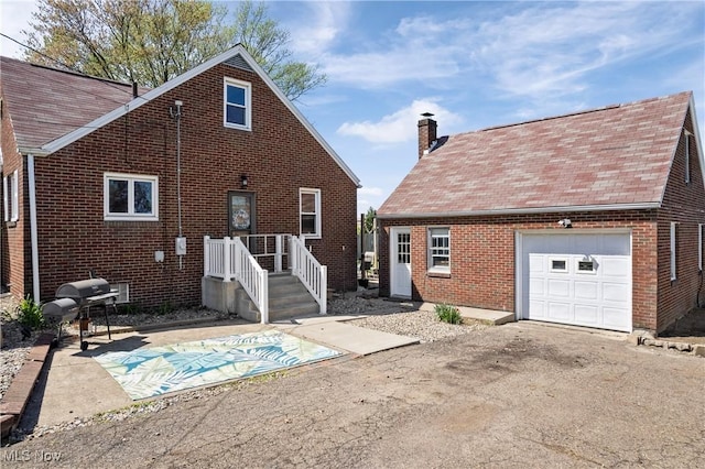 back of property featuring a garage, roof with shingles, a chimney, brick siding, and driveway